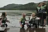 Flere barn ved en brygge ved vannet og to voksne ved rekkverket.