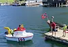 Liten hvit og rød båt med norsk flagg ved en brygge, med to personer på kaien og havn i bakgrunnen.