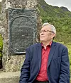 En mann i blå jakke og rød skjorte står ved en minnestein foran bratte, grønne fjell.
