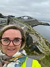 Kvinne i refleksjakke tar selfie ved en kystvei med bru og hav i bakgrunnen.