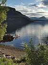 Fjell og skog rundt en stille innsjø med liten brygge og steinete strand i forgrunnen.