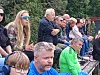 Flere personer sitter på en utendørs tribune og følger med på noe utenfor bildet.