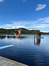 Person i luften over stille innsjø nær en brygge og et lite tårn, med skog og blå himmel bak.
