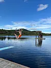 Person i luften over stille innsjø nær en brygge og et lite tårn, med skog og blå himmel bak.