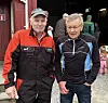 To eldre menn står foran en rød bygning med andre personer i bakgrunnen.