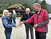 En mann i rød jakke serverer mat til en kvinne på en grusplass foran en hytte.