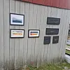 Flere innrammede bilder og små tavler henger på en lys trevegg utendørs.