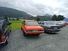 Eldre biler parkert på en grusplass ved en fjord med fjell og skyet himmel.