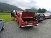 Rød og svart klassisk bil parkert på grus med åpne motorrom foran fjell og vann.