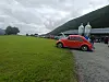 Rød folkevogn parkert på grus ved grønt fjell, med andre biler og mennesker i bakgrunnen.