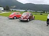 To røde Volkswagen Boble står på en grusplass ved en innsjø med fjell og skog i bakgrunnen.