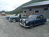 To klassiske biler står parkert på en grusplass ved en bygning med fjell i bakgrunnen.