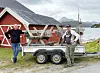 To menn ved en henger foran en rød sjøbod med fjell og fjord i bakgrunnen.