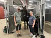 En hest står mellom to personer i en stallgang innendørs.