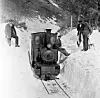 Lite damplokomotiv på snøfylte skinner med menn som står ved siden av i et vinterlandskap.