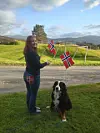 En kvinne holder to norske flagg på en gresslette, med en hund ved siden av og fjell i bakgrunnen.
