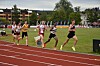 Friidrettsløpere i ulike drakter konkurrerer på en rød løpebane foran tribuner og grønt gress.