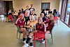 Barn og voksne i håndballdrakter sitter samlet ved små bord i en lys spisesal med røde stoler.