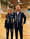 To personer i mørkeblå sportsdrakter står på en innendørs håndballbane.