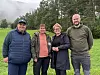 Fire personer står sammen utendørs foran en gressplen med trær, fjell og skodde bak.