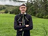 En ung mann står foran en eng med skog og fjell i bakgrunnen.