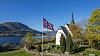 Hvit kirke ved fjord med norske flagg, trær og fjell i bakgrunnen.