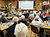 Mange eldre personer sitter i et møtelokale mens en presentasjon vises på lerret foran i rommet.