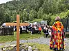 Utendørs samling med kors og en kappekledd person med ryggen til i forgrunnen, stoler og mennesker samlet i et skogsområde.