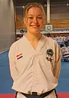 Smilende person i hvit kampsportdrakt står i en hall med blå matte og folk i bakgrunnen.