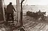 Fisker på brygge ved vannet med båt og flere personer nær land.