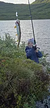 Barn med fiskestang ved en innsjø, med en nyfanget fisk hengende fra snøret.