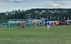 Fotballbane med mål, spillere, tilskuere og telt foran en boligbakgrunn.