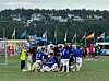 Fotballspillere i blått og hvitt samles foran målet på en bane med flagg og hus i bakgrunnen.