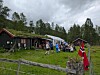 Mennesker står samlet utenfor svarte trehus på en gressbakke med norsk flagg foran.