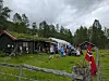 Mennesker står samlet utenfor svarte trehus på en gressbakke med norsk flagg foran.