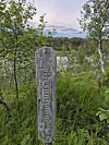 Værslitt trepåle med tekst i grønn vegetasjon ved vann og skog.