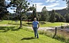 Mann står på en grønn eng ved en vei med trær og åser i bakgrunnen.