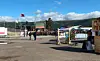 Utendørs torg med boder, telt og folk, omgitt av flagg og fjell under blå himmel.