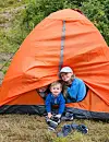To barn sitter ved inngangen til et oransje telt på gresset med grønn skog i bakgrunnen.