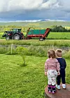 To små barn står på en sti og ser på en traktor med tilhenger på et jorde.