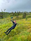 En gutt svinger i en line over en grønn eng med skog og grå himmel i bakgrunnen.