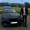 En mann står ved en svart BMW parkert foran et SUTRA-skilt med fjell og åker i bakgrunnen.