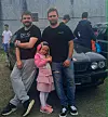Tre personer står foran en mørk BMW på en utendørs parkeringsplass.