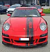 Rød Porsche sportsbil med svart dekor sett forfra, parkert blant andre biler utendørs.