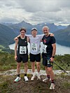 Tre løpere står arm i arm på en fjelltopp med fjellandskap og innsjø bak seg.