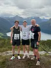 Tre løpere står arm i arm på en fjelltopp med fjellandskap og innsjø bak seg.