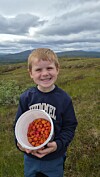 Et smilende barn holder en hvit bøtte med oransje bær ute i et gresskledd landskap.