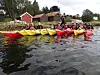Padlere i gule og røde kajakker på vannet ved en steinete kyst med røde hus i bakgrunnen.