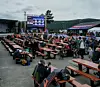 Publikum sitter ved bord foran en scene utendørs med fjell og overskyet himmel i bakgrunnen.