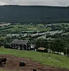 Grønt gårdslandskap med mørk bygning, beitedyr og skogkledd ås under overskyet himmel.